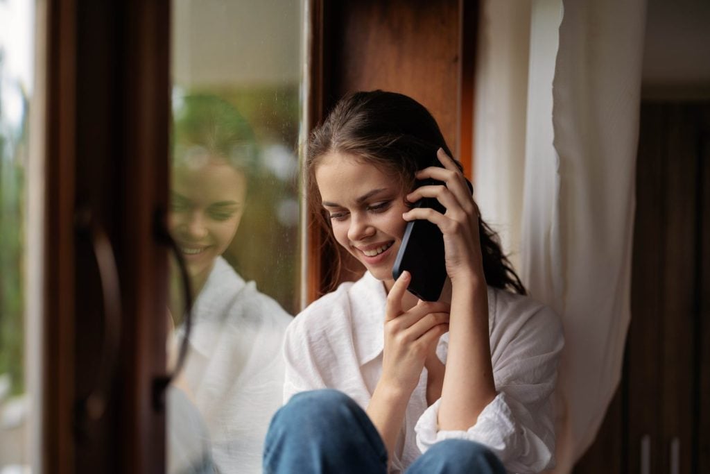 woman sitting on a wooden window sill by the window with a phone in hand