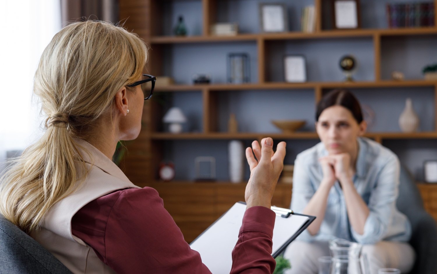 Over shoulder view of female psychologist sitting in armchair, talking with upset woman patient
